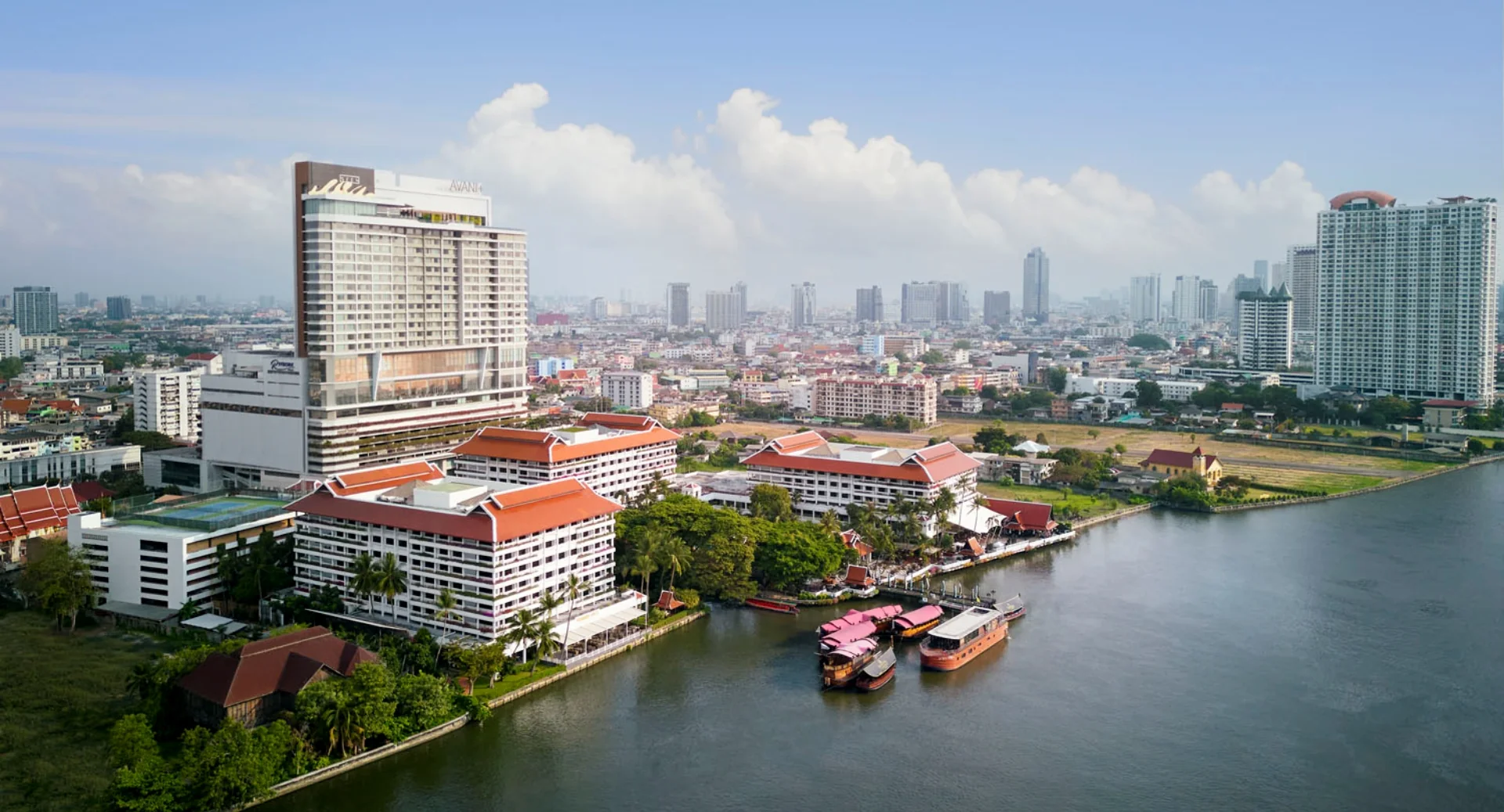Der Turm des Avani+ Riverside mit AVANI-Schriftzug hinter den separaten Anantara-Gebäuden mit roten Dächern am Chao Phraya.