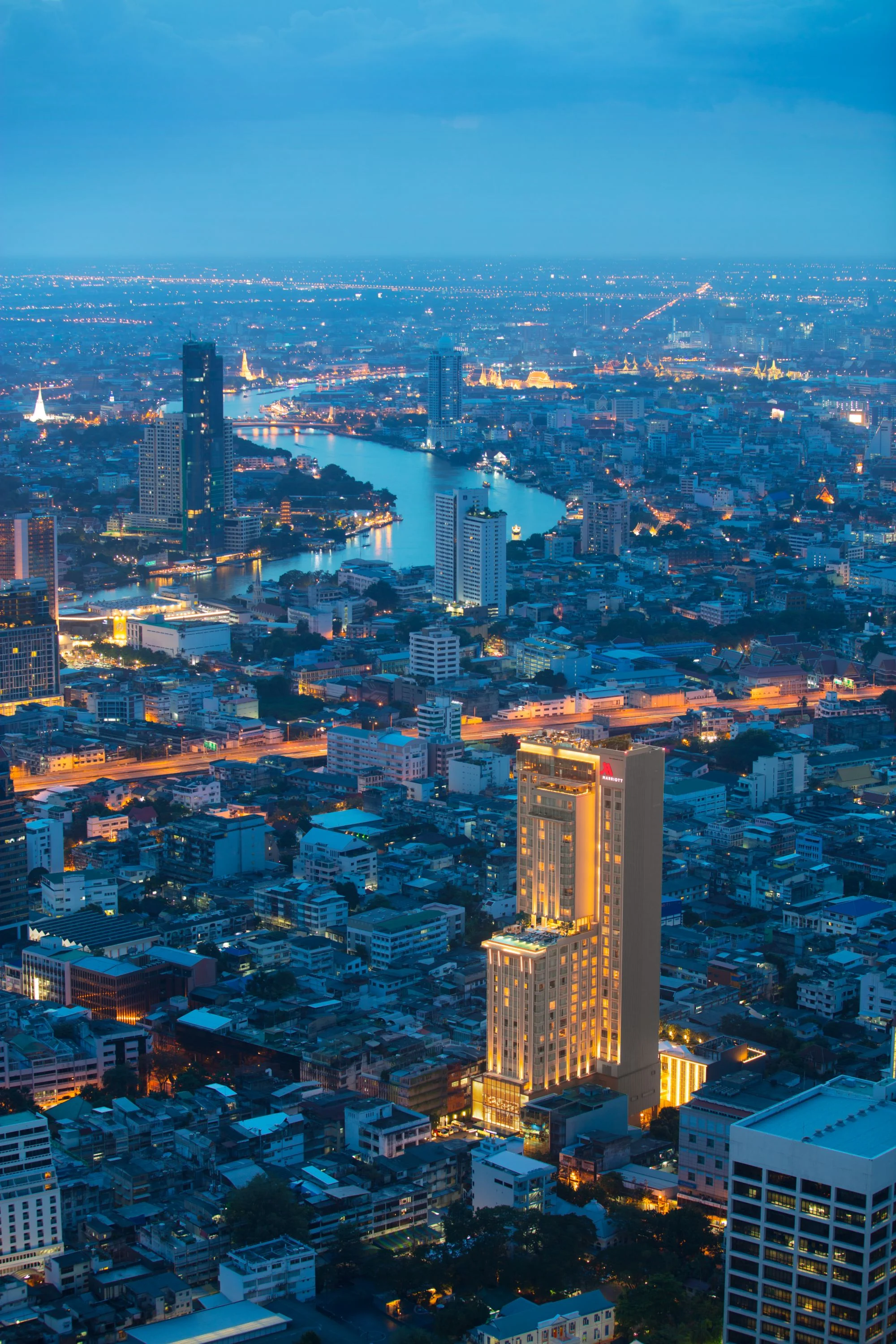 Bangkok Marriott Hotel The Surawongse’s illuminated hotel tower amid Bangkok at blue hour, with the Chao Phraya River in the distance