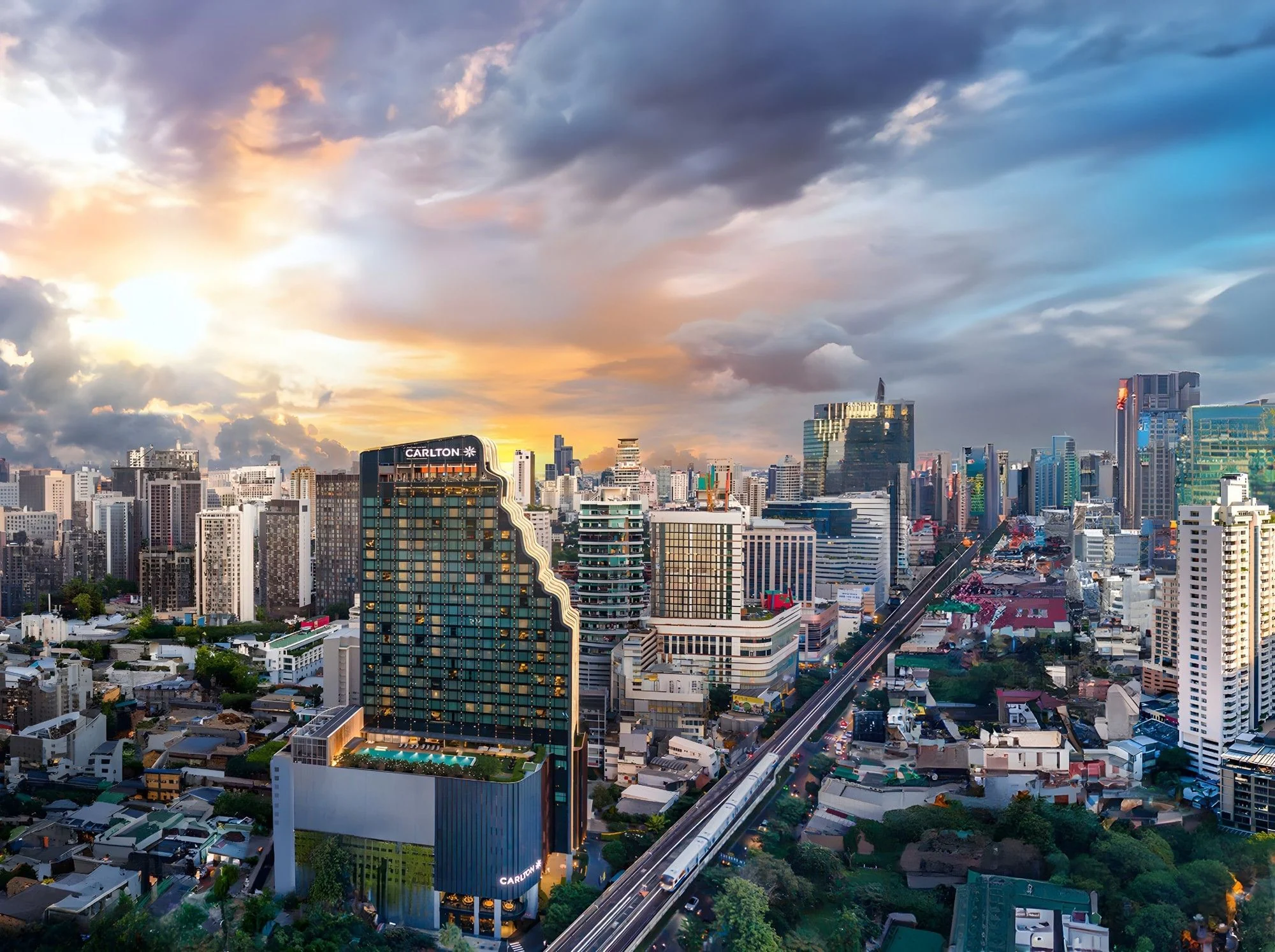 Hotelturm des Carlton Hotel Bangkok Sukhumvit mit Namensschild neben der Sukhumvit Road und der Hochbahn bei Sonnenuntergang
