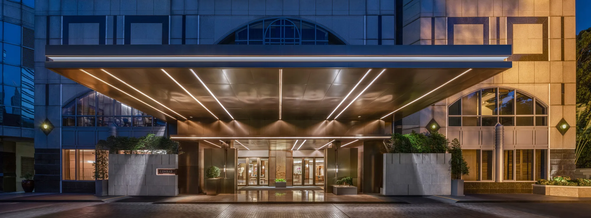 Eingang des Conrad Bangkok mit breitem beleuchtetem Vordach, Glastüren und Hotelnamen in der Abenddämmerung