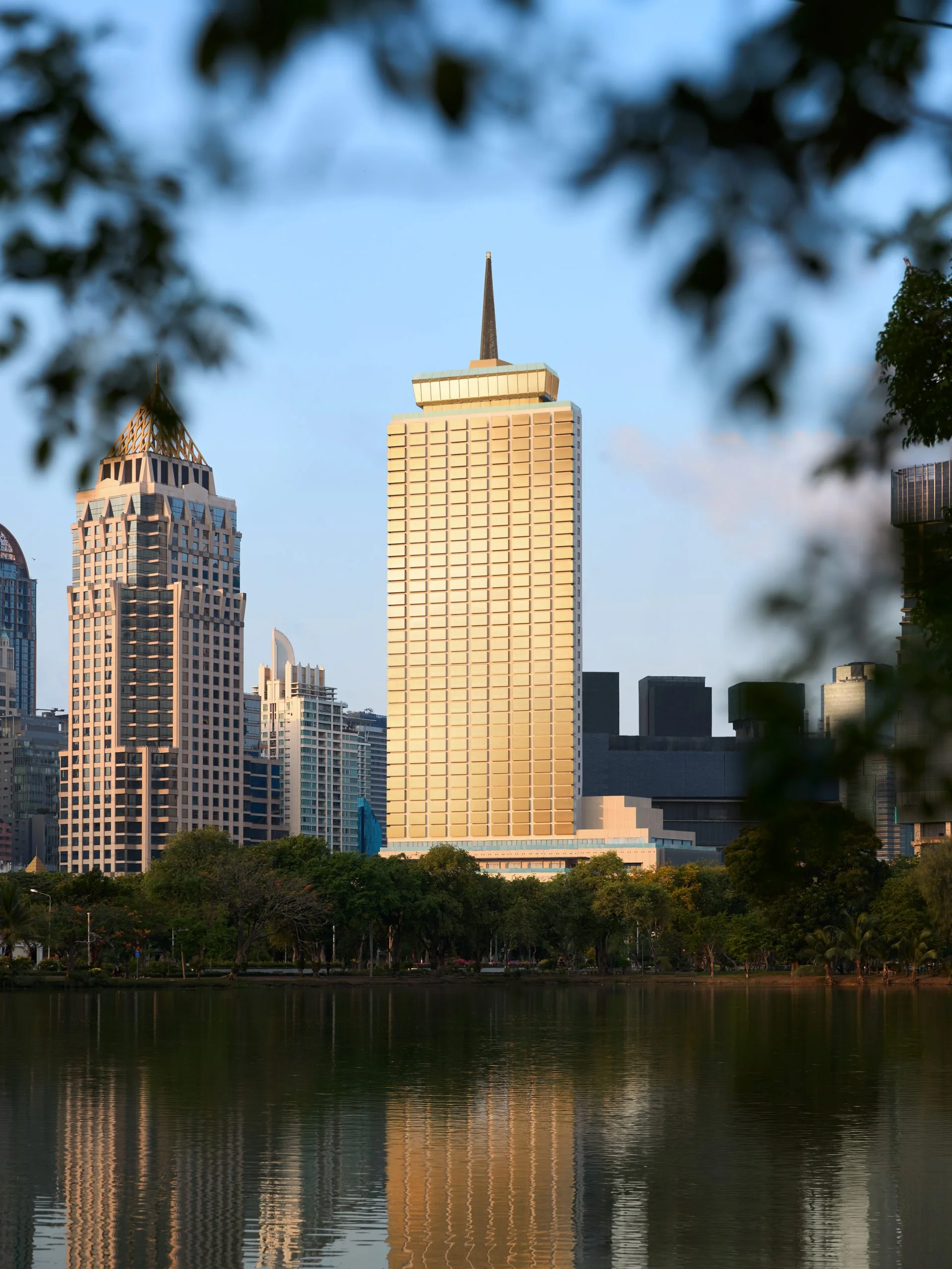 Der goldfarbene Turm des neu errichteten Dusit Thani Bangkok mit seiner Spitze über dem Lumpini-Park, bei Tageslicht vom anderen Seeufer aus gesehen