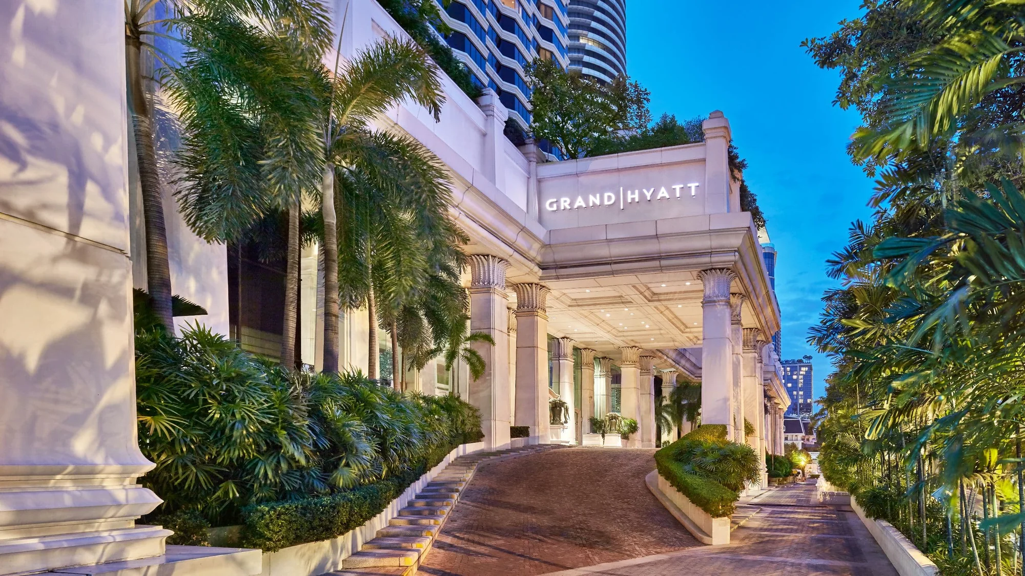 Beleuchtetes Eingangsdach des Grand Hyatt Erawan Bangkok mit Grand-Hyatt-Schriftzug über der ansteigenden Vorfahrt und Palmen in der Dämmerung