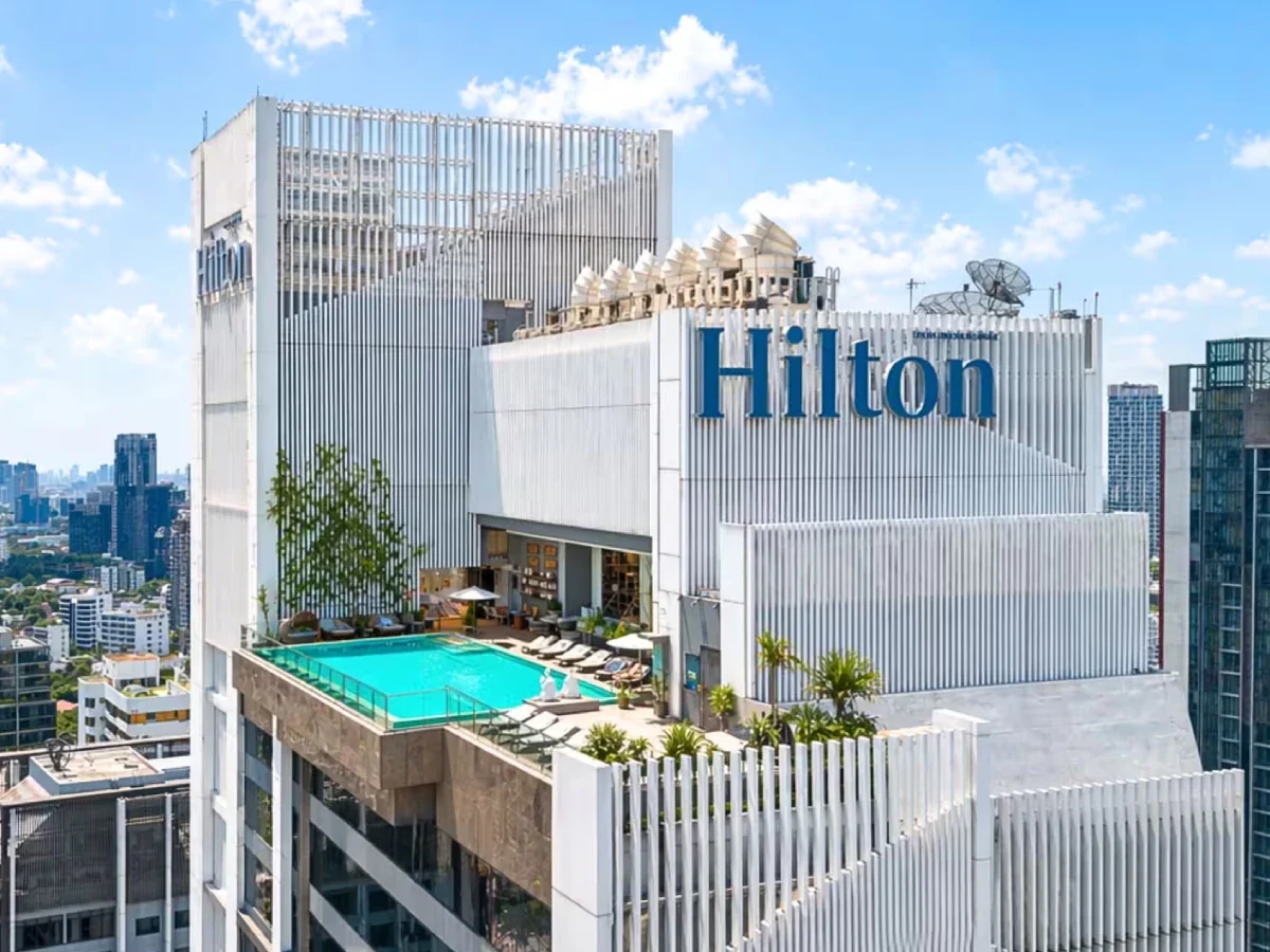 Hilton Sukhumvit Bangkok’s white hotel tower and large Hilton sign in daylight, with its rooftop terrace visible