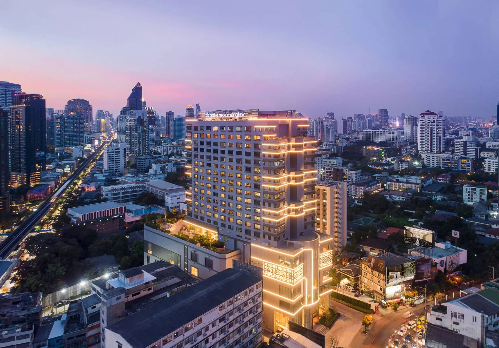 Der beleuchtete Turm des Hotel Nikko Bangkok mit seinem Hotelschild auf dem Dach über Thonglor in der Abenddämmerung