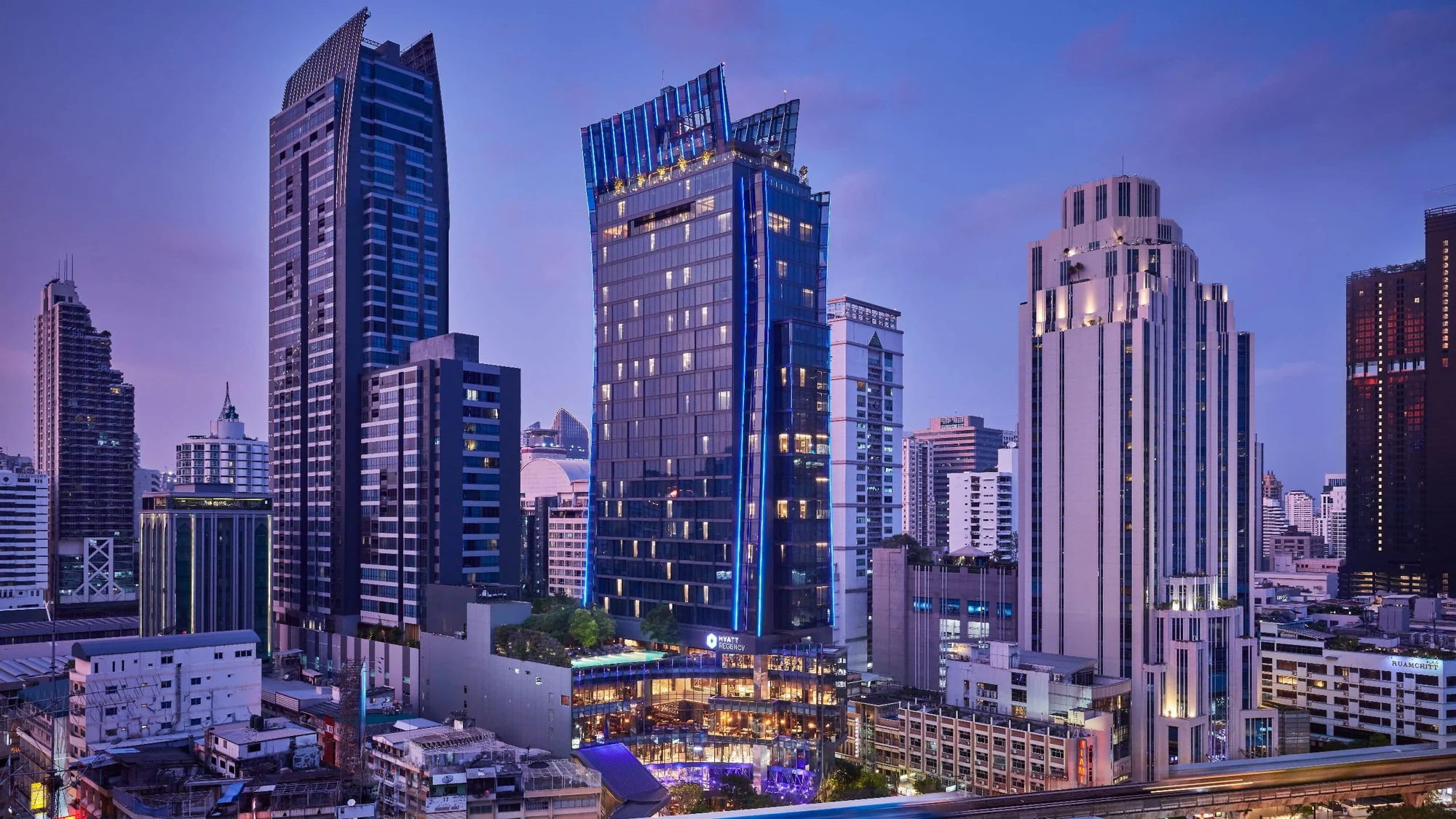 Der beleuchtete Turm des Hyatt Regency Bangkok Sukhumvit mit Hotelschriftzug an der Sukhumvit Road in der Dämmerung.
