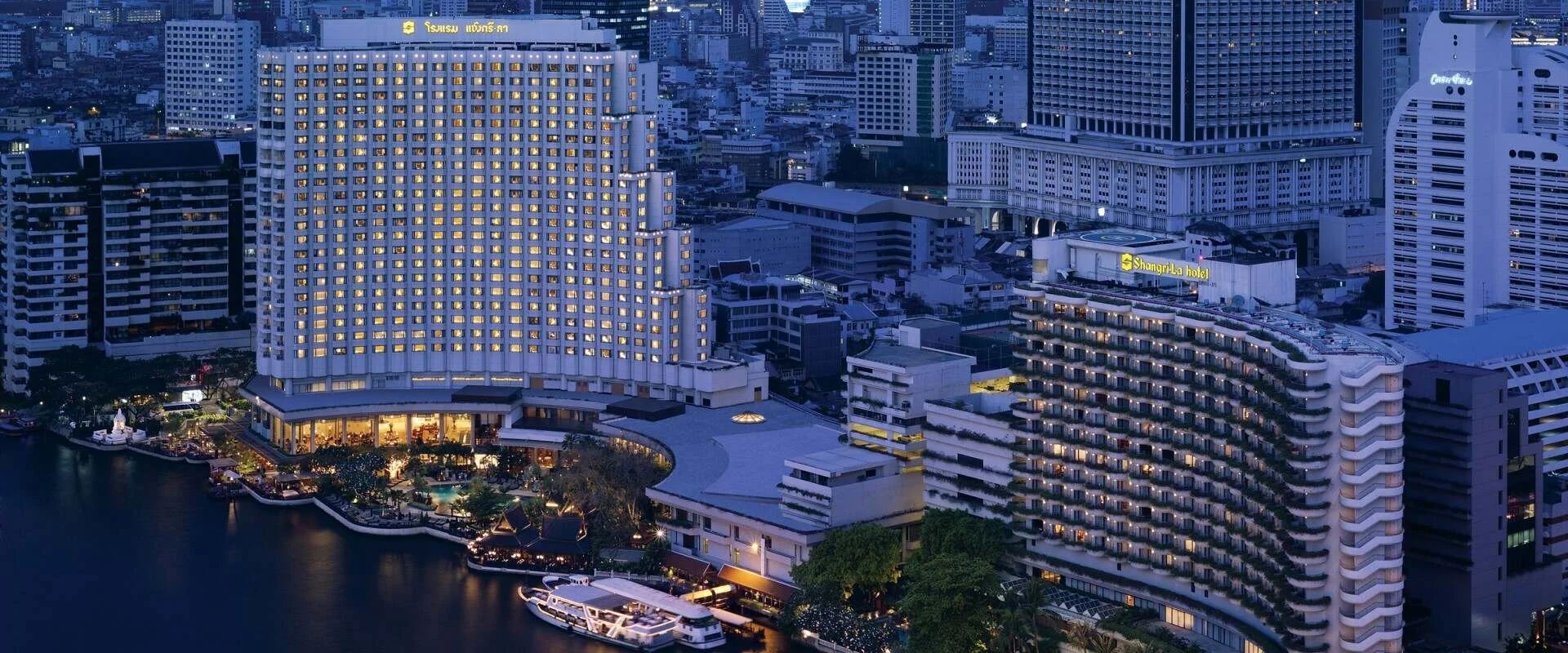 Die beiden beleuchteten Flügel des Shangri-La Bangkok mit ihren Hotelschildern am Chao Phraya in der Abenddämmerung
