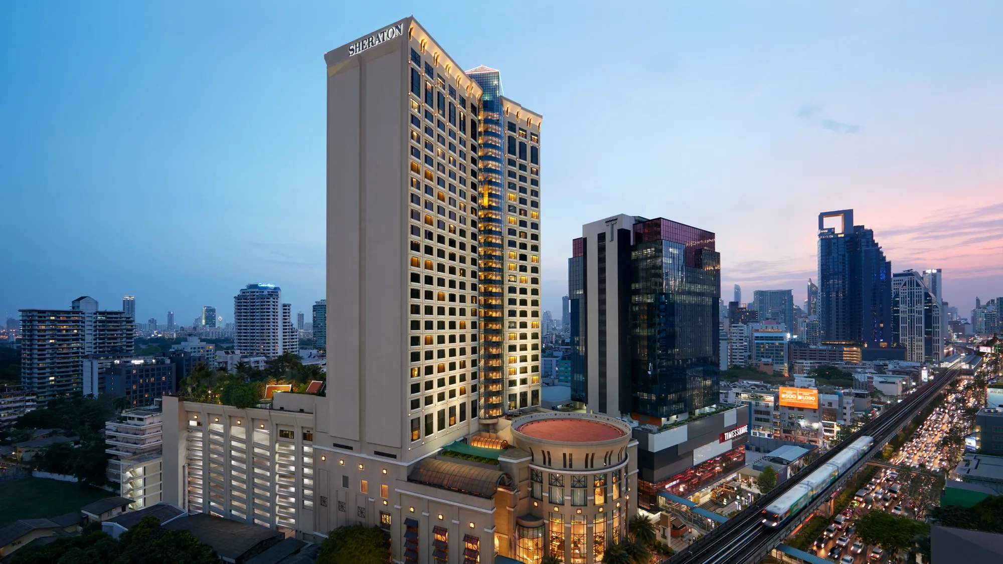 Turm des Sheraton Grande Sukhumvit mit beleuchtetem Hotelnamen und abgerundetem Eingangsgebäude an der Sukhumvit Road in der Dämmerung