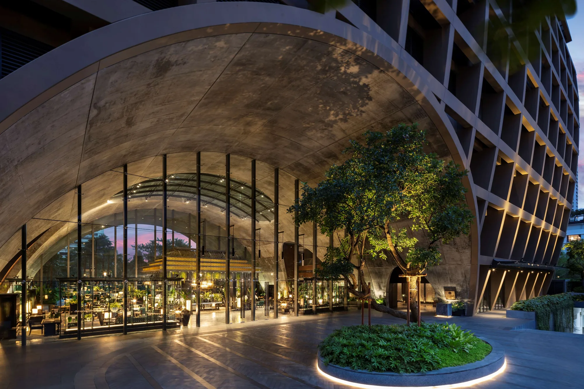 Sindhorn Kempinski Hotel Bangkok mit weit gespanntem Eingangsbogen und beleuchteter Glaslobby in der Abenddämmerung