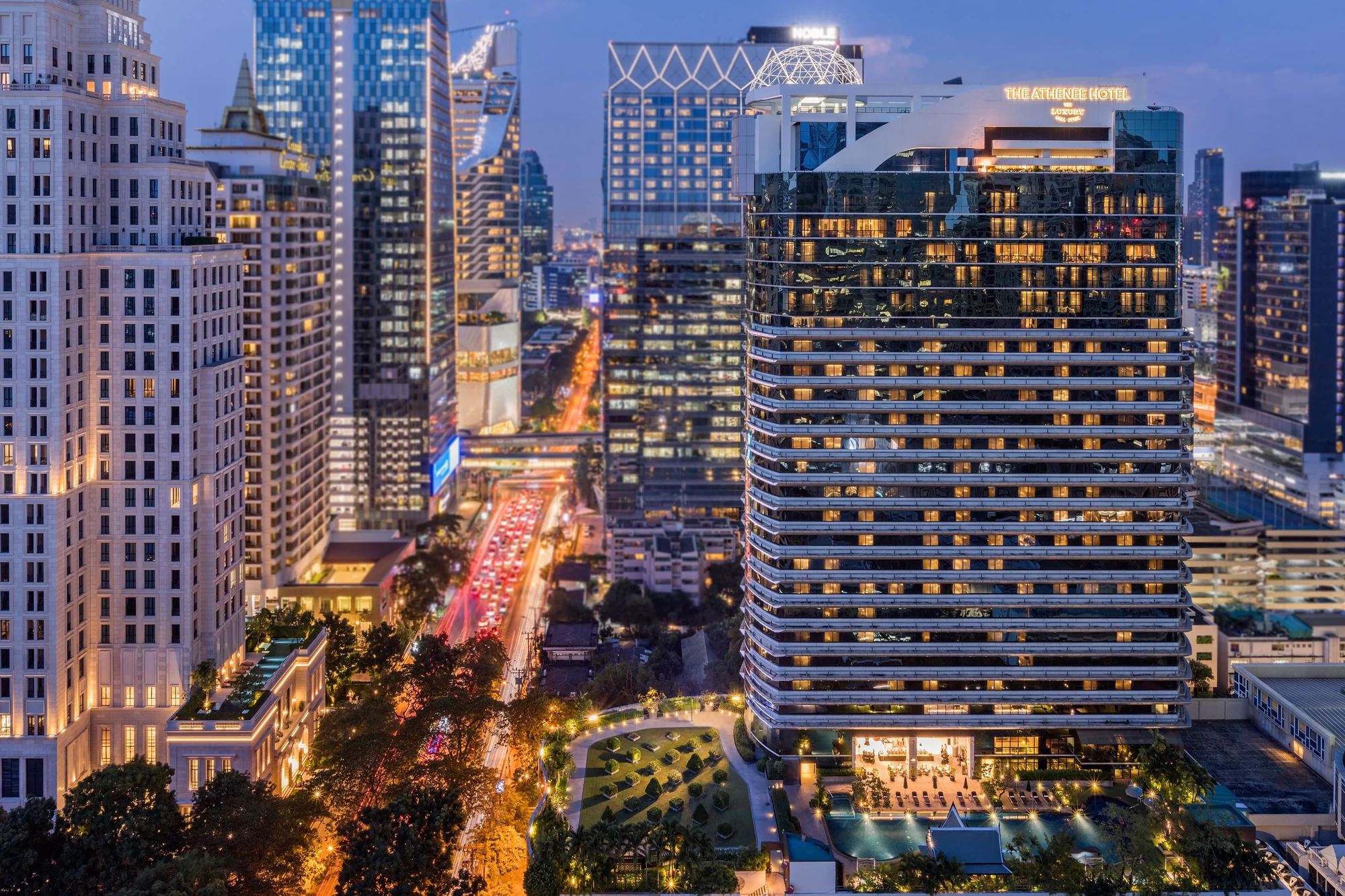 Der beleuchtete Turm des The Athenee Hotel Bangkok mit dem Hotelschild über der Wireless Road in der Abenddämmerung