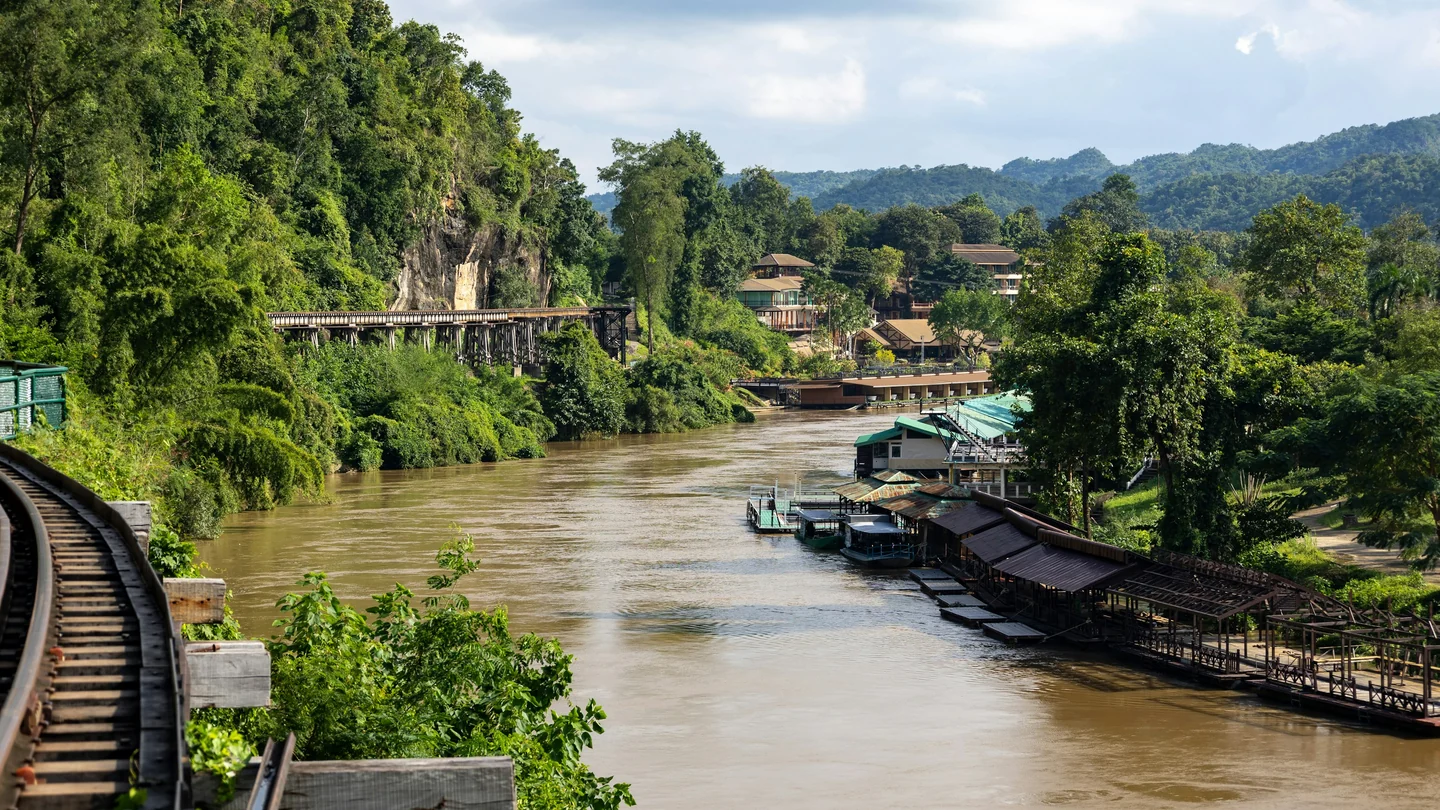A river beside forested cliffs, the wooden railway and riverside buildings at Tham Krasae in Sai Yok